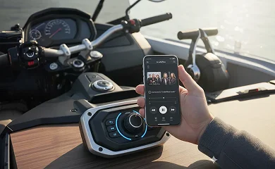 Hand holds a smartphone over a boat or motorcycle console with audio controls and speaker knob visible.